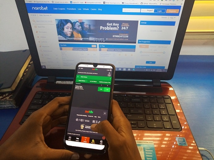 An African's hands holding a phone in front of a laptop, both open to sports betting sites.