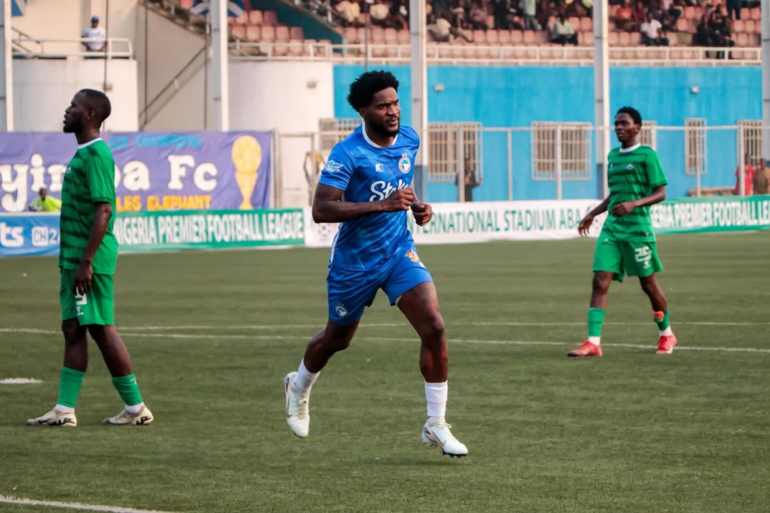 Brown Ideye wearing a blue jersey running and celebrating a goal, flanked by two opponents in green jerseys.