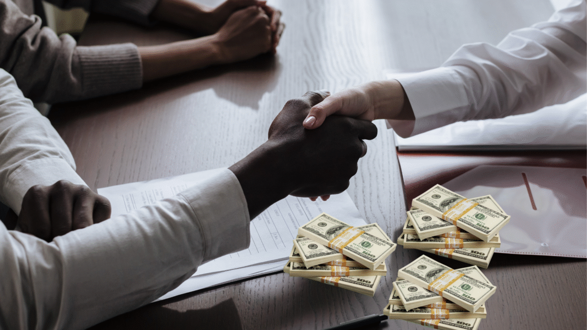 black and white hands locked in a handshake over a table with wads of money