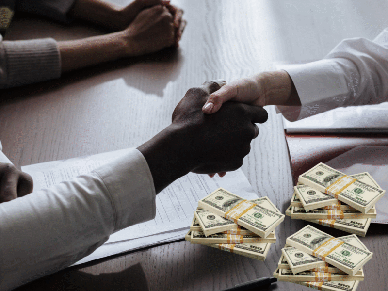 black and white hands locked in a handshake over a table with wads of money